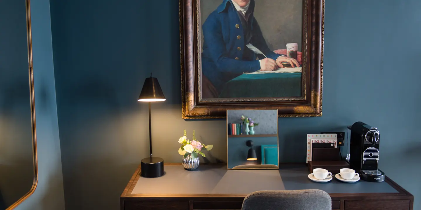 A painting depicting a man seated at a desk framed above a wooden desk.