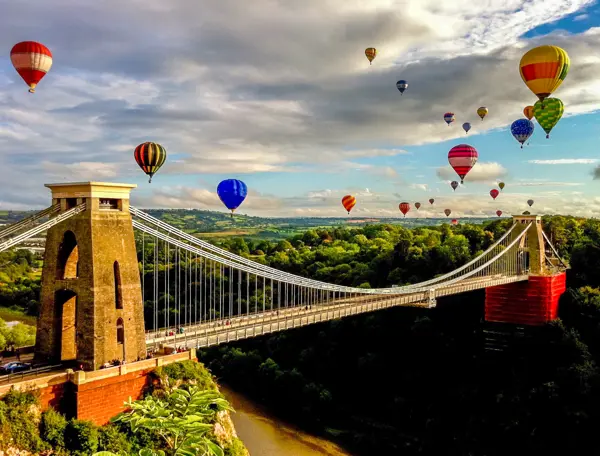 Numerous hot air balloons soar above a bridge.