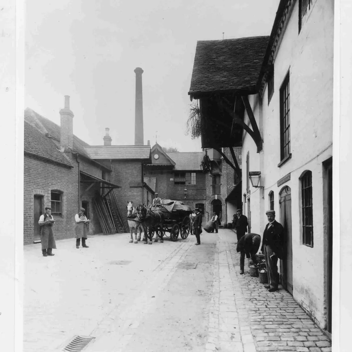 Black‑and‑white historic photograph of the interior of the former Brakspear Brewery, showing a horse‑drawn carriage and seven male workers. One man is rolling a barrel, two are taking notes, and others are posing for the camera.