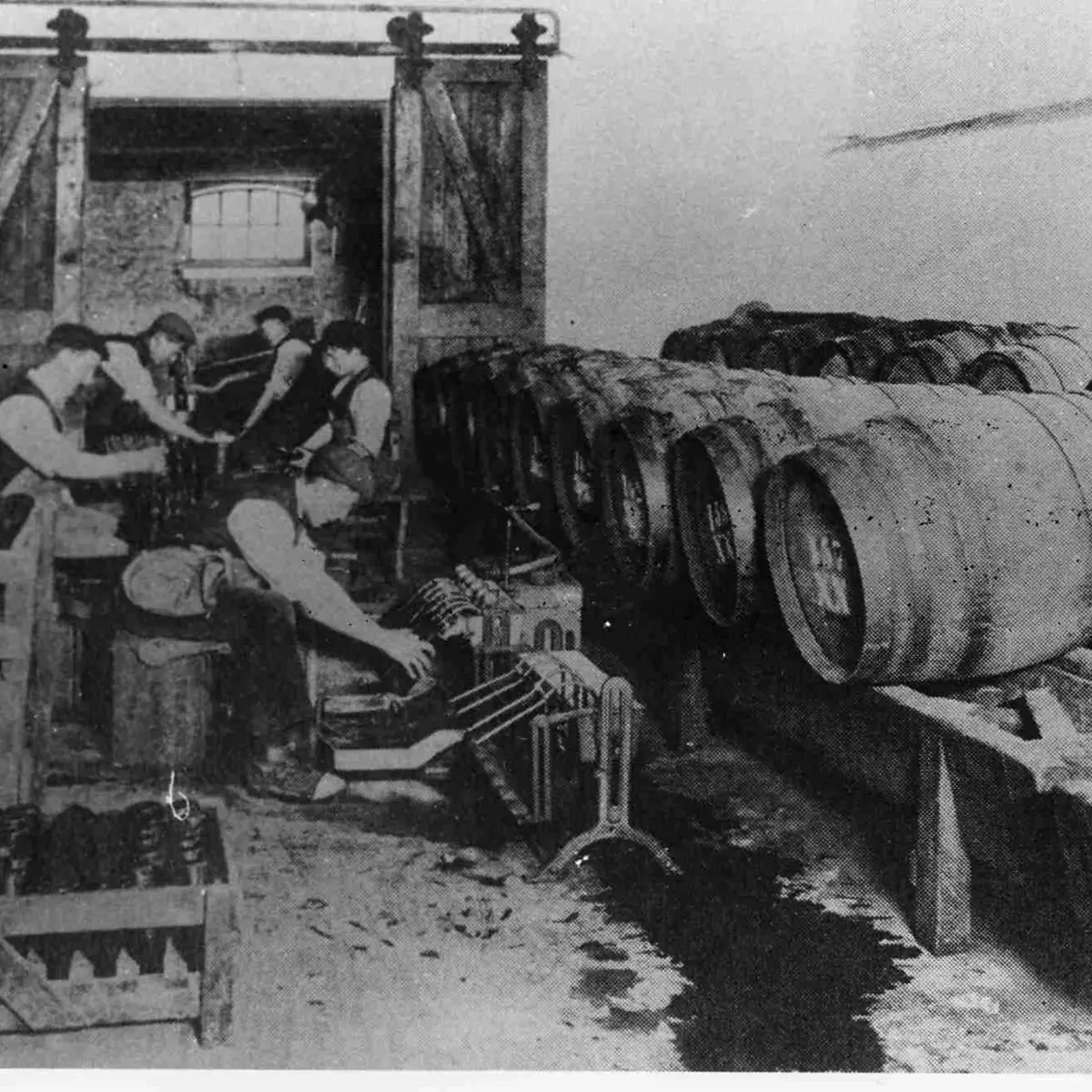 Black‑and‑white historic photograph showing five men working inside the former Brakspear Brewery. Several barrels stand behind them, and bottles are stored on the floor, protected by a low wooden fence.